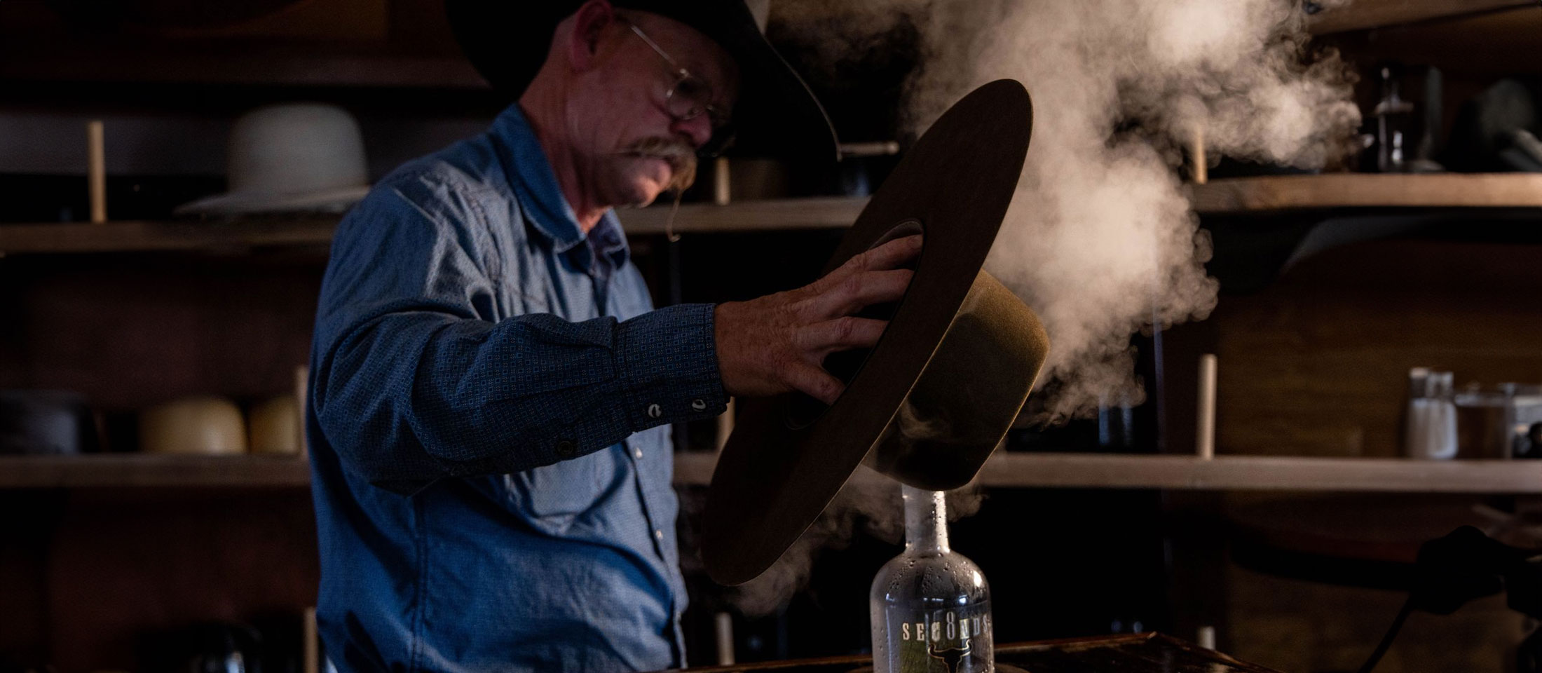 A man, Chaz Mitchell, standing in front of a hat steamer, steaming a brown cowboy hat.